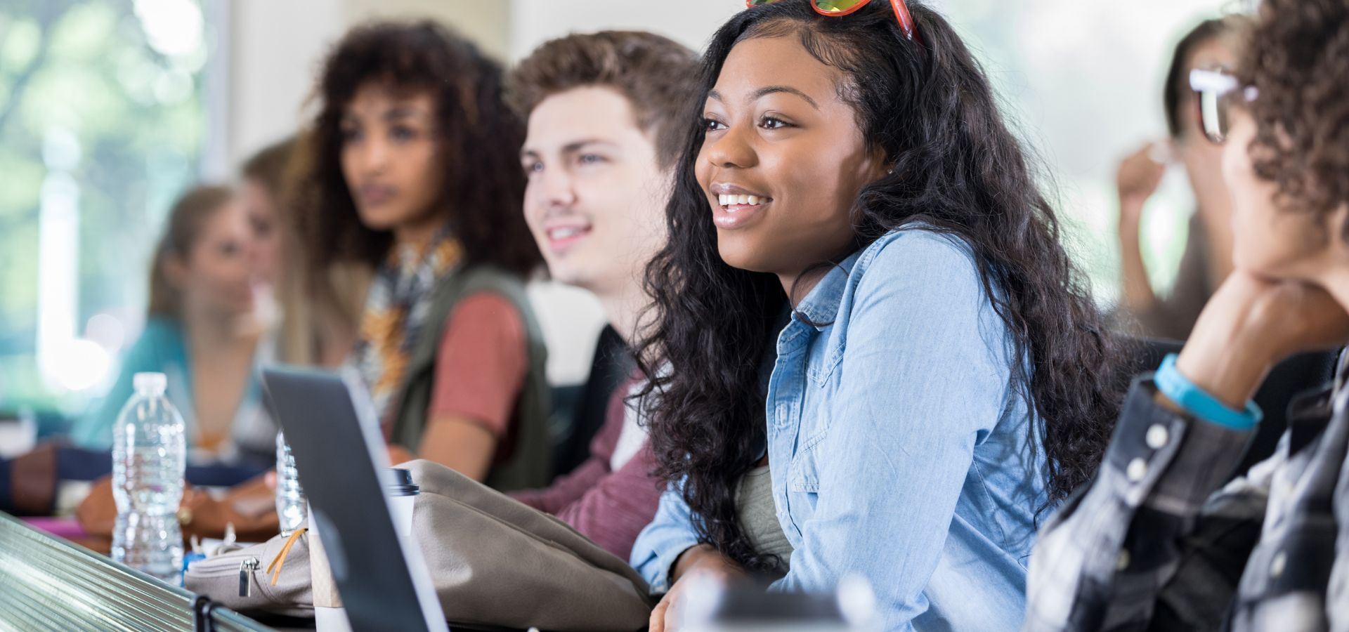 Diverse college students smiling and engaged during a classroom lecture, with a young woman in a denim shirt in the foreground.